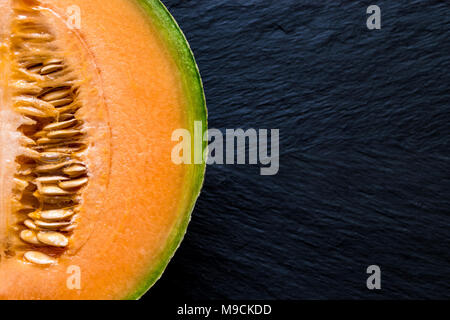 Die Hälfte der Hami melone Frucht auf schwarzem Stein Hintergrund Oberfläche mit freiem Speicherplatz. Hintergrund ist unscharf, liegt der Schwerpunkt auf Melone Stockfoto