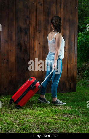 Junge Mädchen von hinten mit Jeans und Turnschuhe schleppen einen roten Koffer, alle über Holz Hintergrund mit Gras Boden Stockfoto