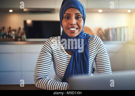 Lächelnden jungen muslimischen Frauen Unternehmer trägt ein Kopftuch, die an einem Notebook arbeitet, während an einem Tisch sitzen in ihrer Küche Stockfoto