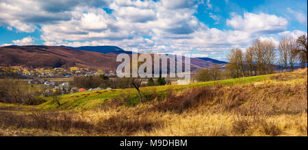 Panorama der bergigen ländlichen Gegend im Frühling. schöne Landschaft an einem bewölkten Tag. blattlosen Wald über der grünen Wiese und Dorf im DISTAN Stockfoto