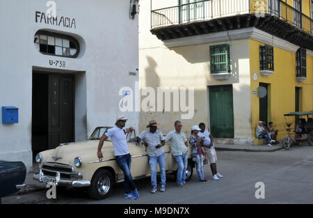 Eine Gruppe von Menschen lehnen an ein klassisches Auto ausserhalb der Apotheke in der Calle Oficios (Straße) in der Altstadt von Havanna, Kuba geparkt. Stockfoto