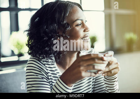 Lächelnde junge afrikanische Frau trinkt eine Tasse Kaffee und schauen durch das Fenster, wenn sie alleine an einem Tisch sitzen in einem Cafe Stockfoto