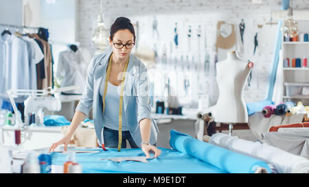 Schöne weibliche Mode, Designer, stützt sich auf ein Stück Gewebe. Sie arbeitet in einem hellen, farbenfrohen Studio voll von verschiedenen Kleidung, Stoffe und Skizzen. Stockfoto