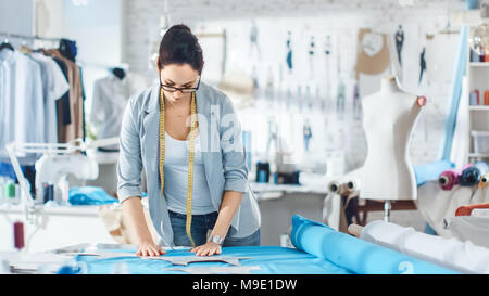 Schöne weibliche Mode, Designer, stützt sich auf ein Stück Gewebe. Sie arbeitet in einem hellen, farbenfrohen Studio voll von verschiedenen Kleidung, Stoffe und Skizzen. Stockfoto