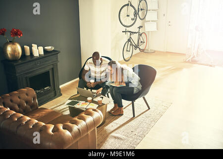 Zwei lächelnde diverse Kollegen sitzen in einem stilvollen, modernen Büro reden und online gemeinsam mit einem Laptop Stockfoto