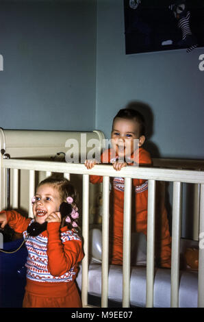 Zwei kleine Kinder vor dem Schlafengehen spielen und tragen in den USA in den 1950er Jahren einen passenden Schlafanzug. Ein kleines Mädchen lacht und hat Haarlocken im Haar, die vorgeben, mit einem Spielzeug-Telefon zu telefonieren. Der Junge ist in einem Kinderbett lachend Stockfoto