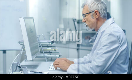 Leitender Arzt der Arbeit in seinem Büro an seinem Desktop-Computer. Auch sein Assistent arbeitet im Hintergrund. Stockfoto
