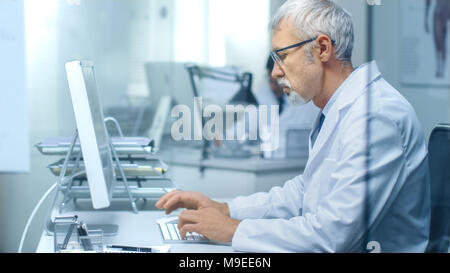 Leitender Arzt der Arbeit in seinem Büro an seinem Desktop-Computer. Auch sein Assistent arbeitet im Hintergrund. Stockfoto