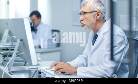 Leitender Arzt der Arbeit in seinem Büro an seinem Desktop-Computer. Auch sein Assistent arbeitet im Hintergrund. Stockfoto