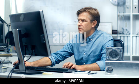 Besetzt Ingenieurbüro. Chief Engineer, der an seinem Schreibtisch, einige technische Komponenten liegen auf seinem Schreibtisch. Sein Assistent arbeitet an seinem Schreibtisch Stockfoto