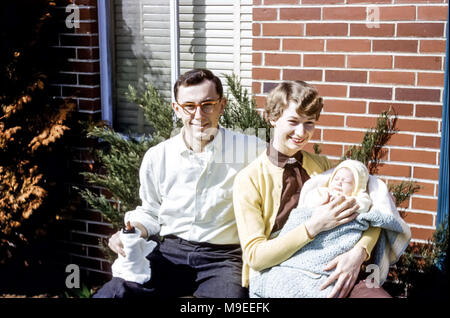 Junges Paar sitzt in Sonnenschein im Freien lächelnd. Die junge Frau hält ein neugeborenes Mädchen in einem Schal gewickelt und der junge Mann hält eine Babyflasche, USA in den 1950er Jahren Stockfoto