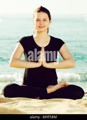 Fitness Woman in Black T-Shirt sitzt und Yoga am Strand. Stockfoto