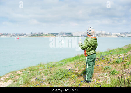 Little Boy in Jacke und Hut auf dem Grasbewachsenen grünen Hang hält gelbe Blume in der Hand und schaut in Richtung Meer mit Segelbooten im kalten Frühling. Stockfoto