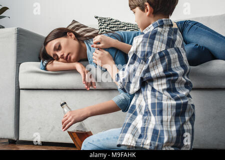 Junge und Mutter Alkoholiker zu Hause Sohn aufwachen Mama schlafen auf dem Sofa betrunken Holding whiskey Flasche close-up Stockfoto