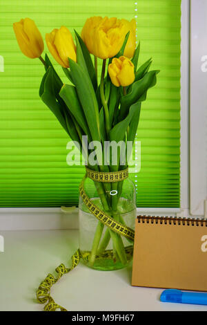 Gelbe Tulpen stehen in Glas Glas auf der Fensterbank. Massband umwickelt ist. Eine leere Notepad für Notes steht neben Marker Stockfoto