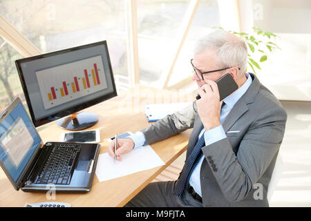 Hohe Aufnahme von Senior Investment Geschäftsmann tragen Anzug, während im Büro Schreibtisch vor dem Laptop sitzen und zum Telefonieren. Stockfoto