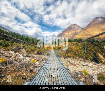 Suspension Bridge und schönen Berge des Himalaja bei Sonnenuntergang im Sommer. Landschaft mit Metal Bridge, Steine, grüne Bäume, hohen Bergen und blauen Clo Stockfoto