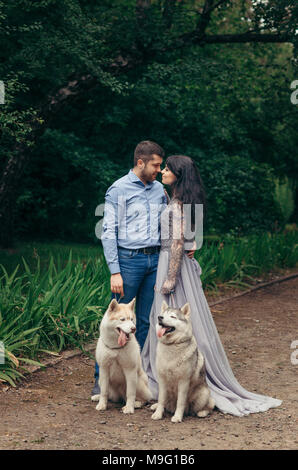 In voller Länge Porträt des Glücklichen adorable elegantes Paar mit zwei Schlittenhunde während ihrer Wanderung im Frühjahr Park. Frau im langen Abendkleid reibt Nasen mit ihrem Geliebten. Stockfoto