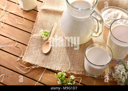 Gläser und Kanne mit Kuhmilch auf sackleinen Tischdecke auf einem Holztisch mit dekorativen Blumen outdoor in der Natur. Horizontale Komposition. Stockfoto