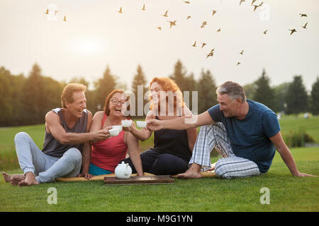 Nach älterer Freunde sitzen und Clink ihre teetassen zusammen. Die Menschen feiern Geburtstag und trinken Kaffee im Park Rasen. Stockfoto