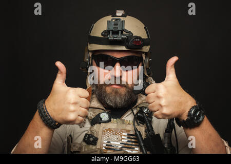 Brutaler Mann in militärischen Wüste einheitliche und Body Armour zeigt zwei Finger auf schwarzen Hintergrund im Studio. Die bärtige Spieler im airsoft Sicherheit Glas Stockfoto