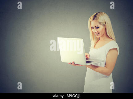 Lässige junge blonde online Einkaufen mit Laptop und glücklich lächelnd auf grauem Hintergrund. Stockfoto