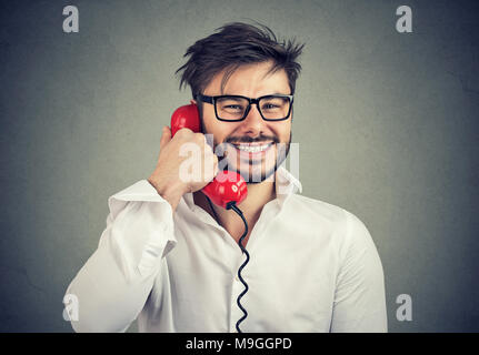 Fröhlicher Mann in Brillen höflich am Telefon sprechen, angenehm und lächelnd an der Kamera auf Grau. Stockfoto
