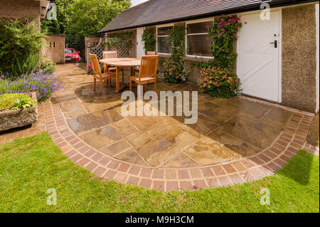 Design & Garten mit gebogenen gepflasterte Terrasse, Rasen, Gitter, hölzernen Tisch & Stühle von Garage des Hauses - West Yorkshire, England, UK. Stockfoto