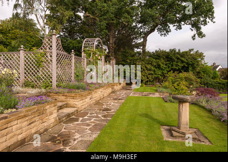 Pfad, blühende Pflanzen auf Grenze, Rasen, Stein, Wand, Holz- Gitter arch und Bildschirm - Schöne, entworfen, Landschaftsgarten, Garten - West Yorkshire, England. Stockfoto