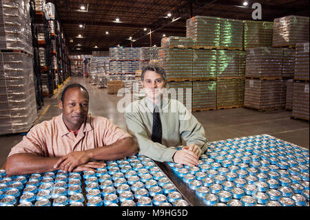 Porträt eines afrikanischen amerikanischen männlichen Lager Vorarbeiter und einem kaukasischen männliche Management Person in ein Lager voller Dosen aromatisiertes Wasser auf der sie gespeichert sind Stockfoto