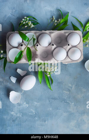 Huhn Eier, ganze und gebrochen, in einem Papier Verpackungen auf einen konkreten Hintergrund mit grünen Blättern und Blumen. Eco kochen Konzept mit kopieren. Stockfoto