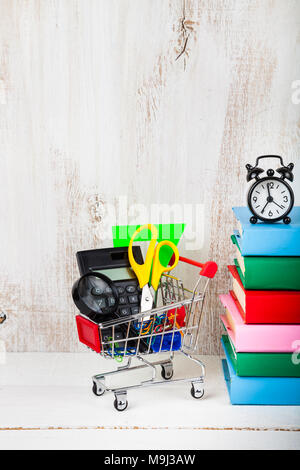 Produkte für Schule in einen Einkaufswagen, Wecker und Bücher auf einer hölzernen Hintergrund. Konzept der Sachen kaufen für die Schule. Zurück zu Schule. Stockfoto