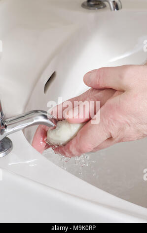 Männliche Hände, hält ein weißer Balken und Seife waschen unter klaren Wasser in Porzellan Waschbecken tippen. Separate heiß und kalt Wasserhähne. Stockfoto