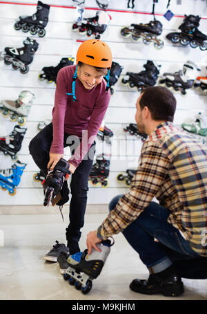 Positive lächelnde Mann Shop Assistant hilft jungen auf Walze zu versuchen - Skates, Sportgeschäft Stockfoto