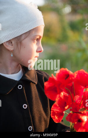 Portrait von niedlichen kleinen Mädchen in weißen Schirmmütze und schwarzen Mantel mit Blumenstrauß aus roten Mohn Blumen im Frühling abends Stockfoto