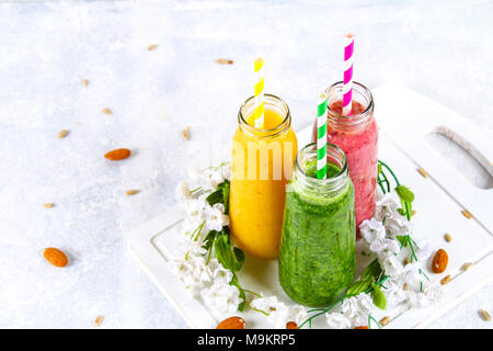 Grün, Gelb, Violett Smoothies im johannisbeere Flaschen, Petersilie, Apfel, Kiwi, Orange auf einem grauen Tabelle mit weißen Blumen Stockfoto