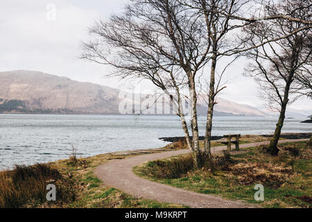 Leere Holzbank durch Loch Eil, Fort William, Schottland, auf einer klaren Frühlingstag. Stockfoto