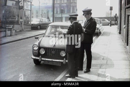 1970 s, historischen, zwei männliche Politessen stehen auf dem Bürgersteig durch ein kleines Auto in einer Seitenstraße, in der Nähe von New Cross Bahnhof in South East London, England geparkt. Stockfoto