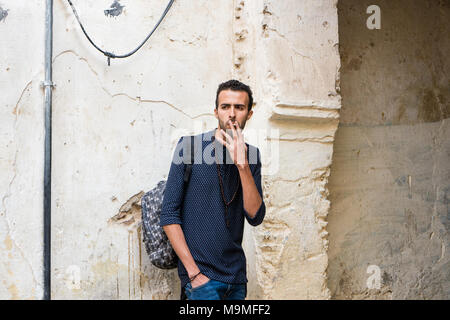 Junge muslimische Mann Rauchen einer Zigarette in Freizeitkleidung stehend durch die zerstörte Wand Stockfoto