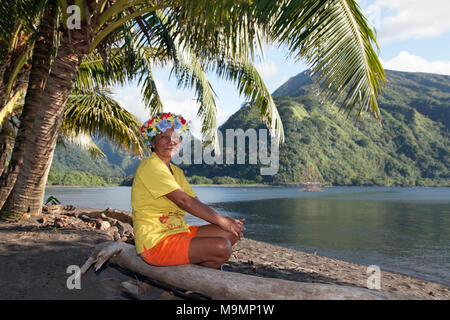 Alte Frau mit Kranz aus Blumen unter Palmen auf Baumstamm sitzend, Küste in der Nähe von Taurita, Tahiti Iti, Gesellschaft Inseln Stockfoto