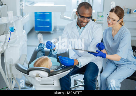 Wichtige Nachricht. Charmante weibliche Krankenschwester zeigt eine wichtige Nachricht auf Tablet zum Zahnarzt während er Untersuchung der Mundhöhle eines kleinen Mädchens Stockfoto