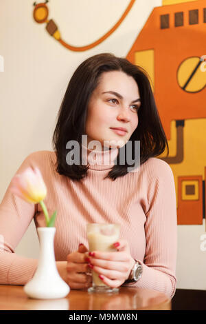 Junge, hübsche Mädchen mit braunem Haar sitzt hinter dem Tisch im Café. Stockfoto