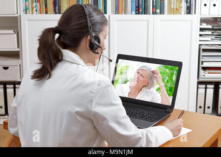 Arzt sitzen am Schreibtisch in Ihrem Büro mit Headset und Laptop, sich Notizen während einer Videokonferenz mit weiblicher Patient leidet unter Haarausfall, Tel. Stockfoto
