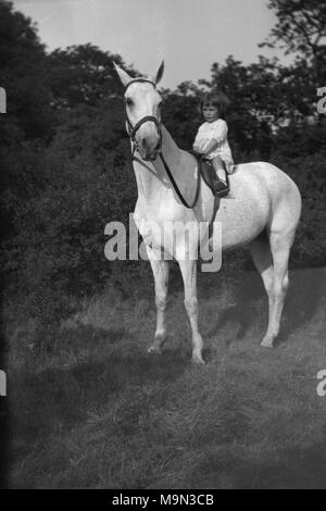 1920s, historische Bild, das ländliche Leben in England, vor der Ankunft auf Masse der Kfz, die meisten jungen Menschen auf dem Land von einem frühen Alter an gelernt, Pferde zu reiten und Hier haben wir ein kleines Kind außerhalb bequem sitzen sehen, die scheinbar furchtlos, auf einem weißen Pferd. Stockfoto