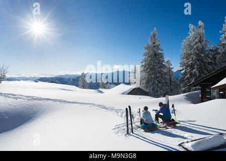 Der Mann und die Frau mit Tourenskiern sitzen auf einer Bank im Schnee am Hubertus Hütte auf dem Berg Breitenstein in der brillanten Winter Sonne, Bayern Stockfoto