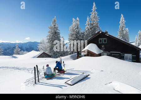 Der Mann und die Frau mit Tourenskiern sitzen auf einer Bank im Schnee am Hubertus Hütte auf dem Berg Breitenstein in der brillanten Winter Sonne, Bayern Stockfoto