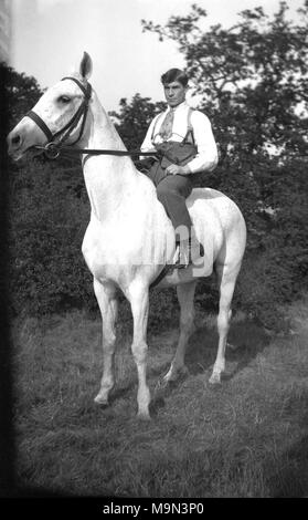 1920s, historische Bild, das ländliche Leben in England, vor der Ankunft auf Masse Der motorcar bedeutete, dass die meisten jungen Leute auf dem Land gelernt, Pferde zu reiten, hier haben wir eine elegant gekleidete junge Mann in einem Hemd, Krawatte und Zahnspangen sitzen bequem auf einem großen weißen Pferd sehen. Stockfoto