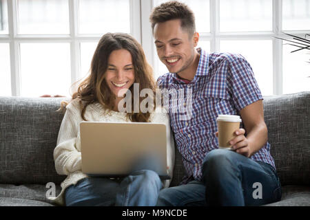 Lächelnden jungen Paar beim Kaffee am Morgen mit Computer sitzen auf dem Sofa zu Hause zusammen, glücklich, Mann und Frau lachend auf der Suche auf dem Laptop Bildschirm hav Stockfoto