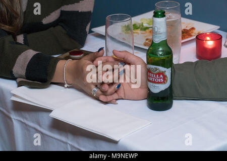 New York City, ca. 2018: Junge Liebe Paar auf Datum halten Hände über Tisch. Leere Teller mit Essen, Kerze, trinken Gläser und Stella Artois Stockfoto