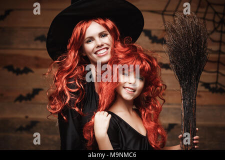 Schönen weißen Mutter und ihre Tochter mit langen roten Haaren in Hexe Kostüme feiern Halloween posiert mit über Fledermäuse und Spinnennetz auf Holz- studio Hintergrund. Stockfoto